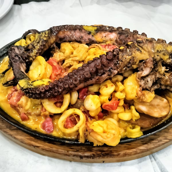 Peruvian grilled octopus with yellow sauce, corn, and red peppers on a sizzling platter.