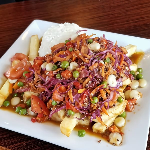 Peruvian poutine with fries, gravy, vegetables, and a dollop of white sauce on a white plate.