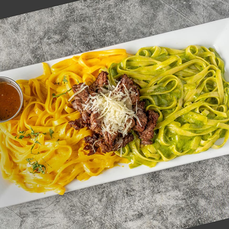 Peruvian green and yellow fettuccine with steak and cheese, served with a side of sauce.