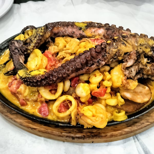 Grilled octopus and seafood in a yellow sauce with corn and red peppers on a sizzling platter.